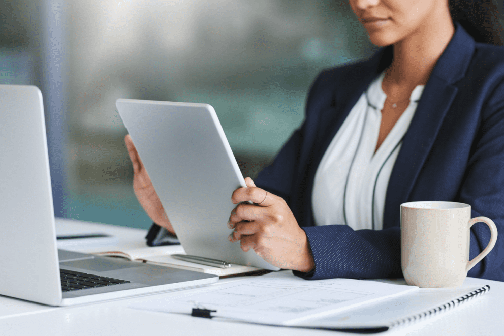 A woman is holding a tablet while her laptop, notes, a coffee cup, and pens sit on a table in front of her.